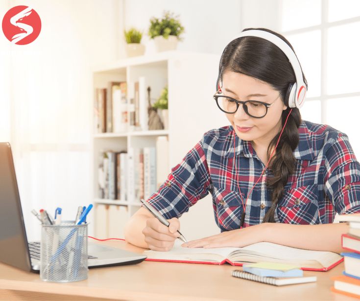 Nữ sinh viên đeo tai nghe, ngồi học tiếng Anh trực tuyến bên máy tính và sách vở